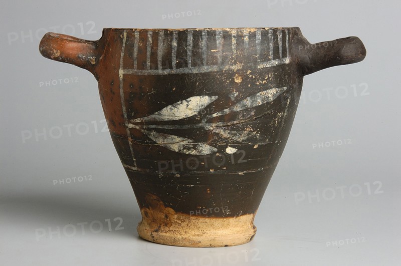 Black-finished skyphos with painted decoration on the rim, white decoration: A = B - laurel branch, IV waC, Etruria, vessel, height 9.7 cm, width 9.2 cm, A ceramic vessel with a tapered body and two short horizontal handles, shown against a pale gray background, featuring a dark brown to black surface with worn lighter areas and painted pale leaf-like shapes on its central body, a band of vertical pale stripes near the rim, and a lighter unglazed beige base that shows signs of wear and discoloration; the handles and upper rim show patches of reddish-brown and flaking, and the overall surface has spots where the dark finish has chipped to reveal lighter underlying material.