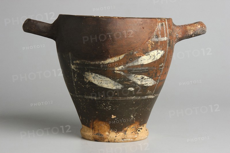 Black-finished skyphos with painted decoration on the finis, white decoration: A = B - laurel branch, IV waC, Etruria, vessel, height 9.7 cm, width 9.2 cm, A ceramic vessel with a wide rim that narrows to a small base, shown from the side with a handle extending from each side; the vessel surface shows worn paint and decorative markings in a lighter color against a darker background, with visible areas of chipped and aged texture; the colors visible include brown, dark brown or black, off-white or cream, and hints of tan and orange on the worn base and rim; the background is a neutral light gray.