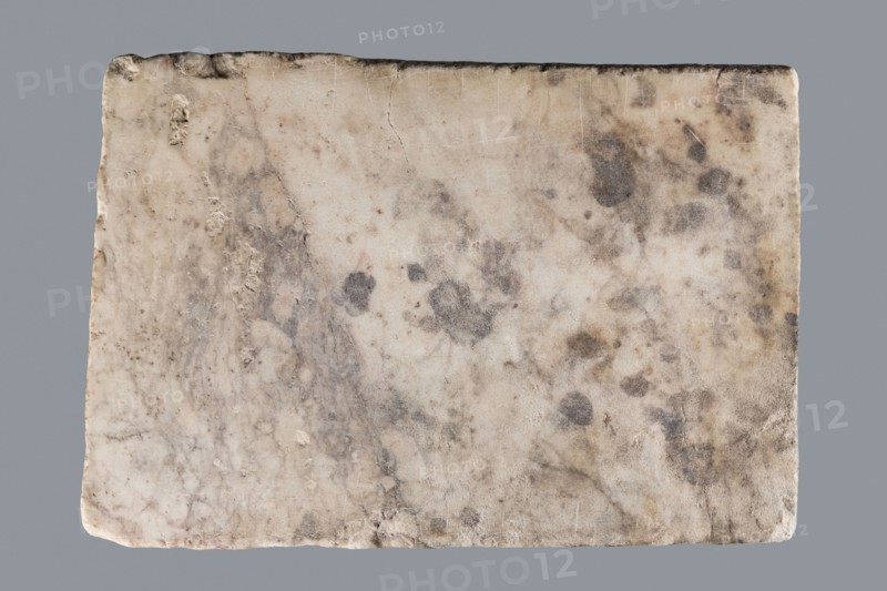 Titulus of the tomb of the Systilians, tombstone inscription in Greek, 2nd–3rd century, Phrygia; material: tombstone; dimensions: height 26.0 cm, width 38.0 cm, depth 4.5 cm., A rectangular stone slab with a rough, worn surface showing a mix of off-white, beige, light tan, gray, and darker gray spots and smudges, with irregular edges and small chips, several rounded darker stains, a few faint veins and streaks across the surface, and an even gray background behind the slab.