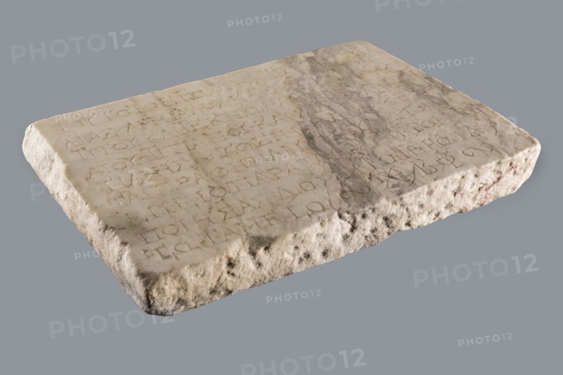 Titulus of the tomb of the Systilians, tombstone inscription in Greek, 2nd–3rd century, Phrygia; tombstone, height 26.0 cm, width 38.0 cm, depth 4.5 cm., A rectangular stone slab rests on a plain background, showing a worn, uneven top surface and rough, eroded edges. The top has shallow carved markings arranged in rows and an irregular darker stain or discoloration across part of it. The stone's colors include shades of beige, off-white, light gray, and areas of darker gray and brownish tones, with some subtle reddish-brown patches along the sides. The texture ranges from smoother, slightly polished areas on the top to coarse, pitted, and crumbly surfaces along the edges.