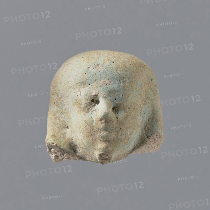 Fragment of a shabti. Fragmentary head of a shabti figurine with indistinct facial features, in a smooth wig. Celadon glaze. Ptolemaic Period, 332–30 BC. Found at El-Kantara. Shabti; height 2.3 cm., A worn pale beige object with patches of light blue and hints of pink that resembles a rounded head with a few small holes where facial features are, a subtle protruding nose shape and an indistinct mouth area, a few rough textured spots and small pits across the surface, and a darker beige irregular base at the bottom, set against a uniform gray background.