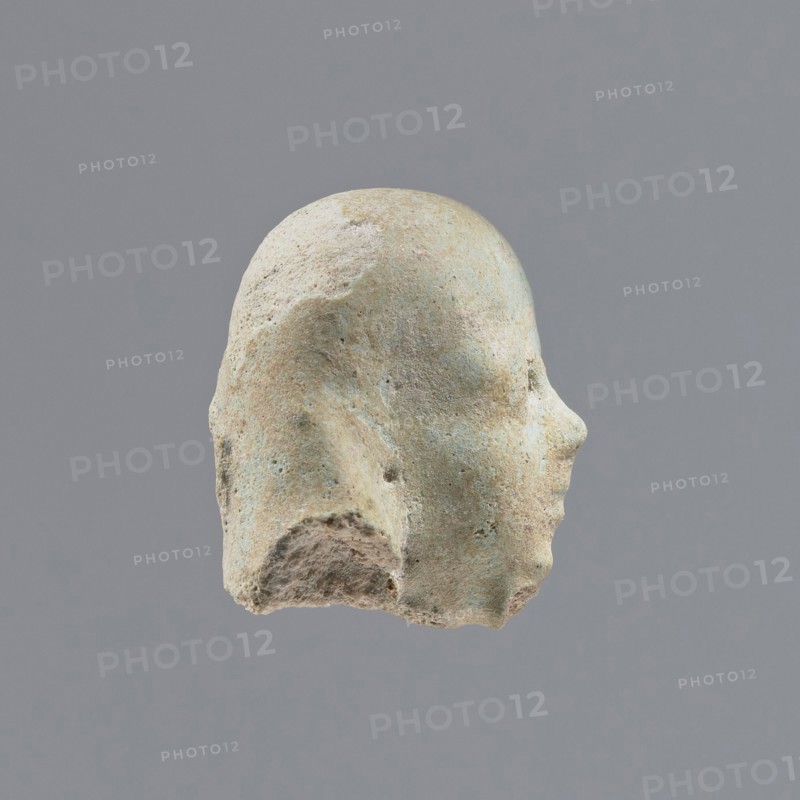 Fragment of a shabti, heads, grave goods, wigs. Head of a shabti figurine with indistinct facial features, in a smooth wig. Celadon glaze. 332–30 BC; Ptolemaic Period. El-Kantara, shabti, height 2.3 cm., A worn sculpted head viewed in profile against a plain gray background, showing a rounded top, a defined nose and forehead, and a rough, eroded lower edge; the surface has a weathered texture with patches of pale beige, light tan, muted green, and hints of bluish-gray, and there are small pits and irregularities across the form.