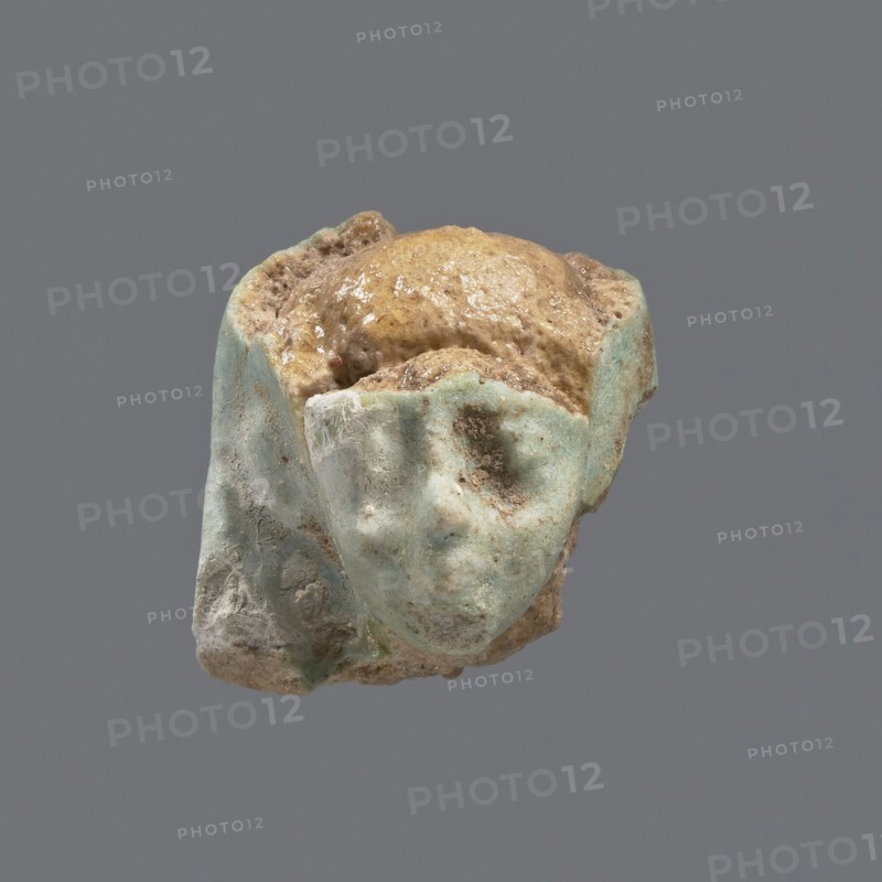 Fragment of a shabti, head of a shabti figurine with carefully rendered facial features in a smooth wig, celadon glaze. Ptolemaic Period, 332–30 BC; found at El-Kantara. Shabti, height 2.4 cm., A small weathered fragment resembling a stylized face sits against a plain gray background, with a worn surface showing a mix of pale turquoise, light blue, beige, tan, and brown tones; the fragment has rounded, smoothed contours with indentations where eye and mouth shapes appear, and a rough, granular texture with some glossy, slightly reflective patches and areas of encrusted debris.