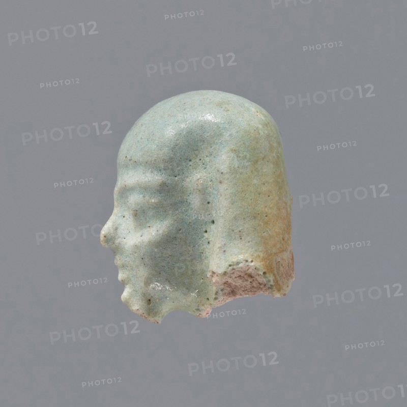 Fragment of a shabti; head of a shabti figurine with carefully rendered facial features, in a smooth wig, with a thin beard. Celadon glaze. Ptolemaic Period, 332–30 BC; El-Kantara. Shabti, height 2.5 cm., A small sculpted head shown in profile with a smooth rounded top and a defined forehead, brow ridge, nose, lips, chin, and a stylized hairstyle that falls down the back of the head, all rendered in a weathered pale green with areas of light blue and touches of beige and tan where the surface is worn, set against a plain medium-gray background.