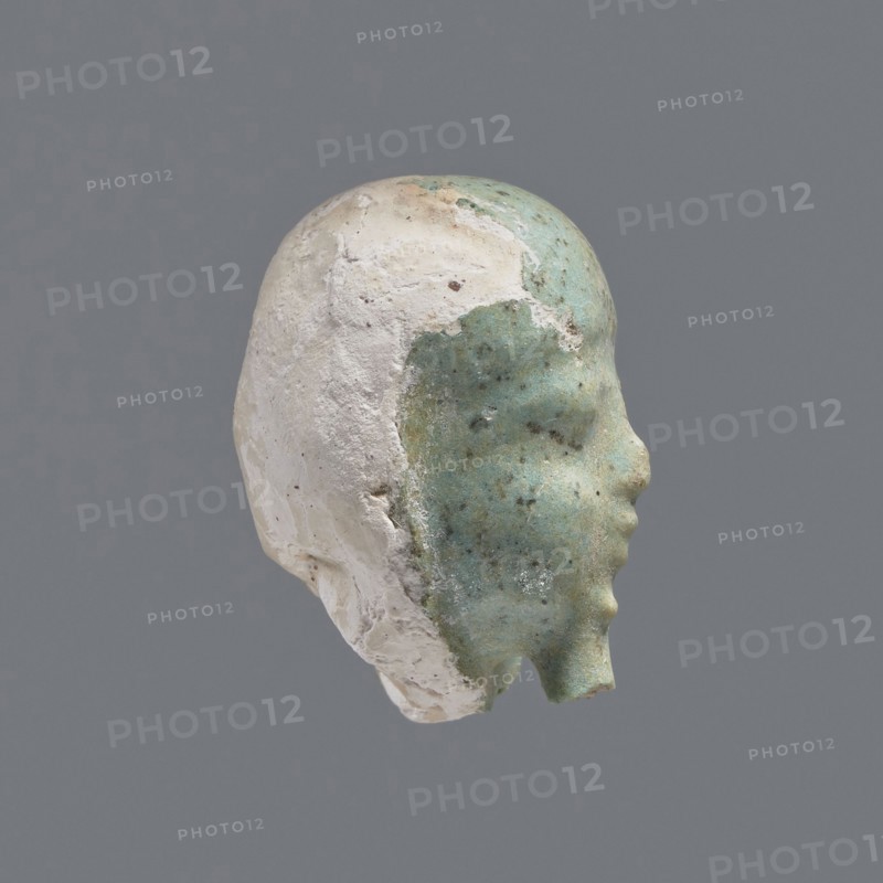 Fragment of a shabti. Head of a shabti figurine with rather carelessly rendered facial features, in a smooth wig and a thin beard. Celadon glaze. 332–30 BC; Ptolemaic Period. Found at El-Kantara. Shabti, height 2.6 cm. Provenance: grave goods; wigs., A sculpted head fragment shown in profile with a smooth face surface that is predominantly greenish-blue mottled with darker speckles and patches, while the rear and top of the head are a rough, chalky white with textured, flaking areas; the background is a uniform medium gray.