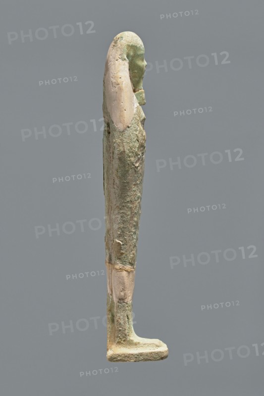 Ushabti, Mummy-shaped figurine, standing on a base with the back resting on a pillar. Arms crossed on the chest, hands indistinctly marked, holding hoes in both hands. Long, smooth three-part wig; ears uncovered. Small, round face with slightly blurred details; long, straight beard adhering to the neck. Painted inscription on the front of the figurine's legs between engraved vertical lines text illegible. Mummy-shaped figurine shabti, Ptolemaic period, 332–30 BC; find location El-Kantara. Height 16.3 cm., A slender standing statue in profile with a long headdress and defined facial features, showing a smooth head and elongated body with arms close to the sides, rendered in a weathered surface combining pale beige and green tones with rough and smoother patches, set against a plain medium gray background.