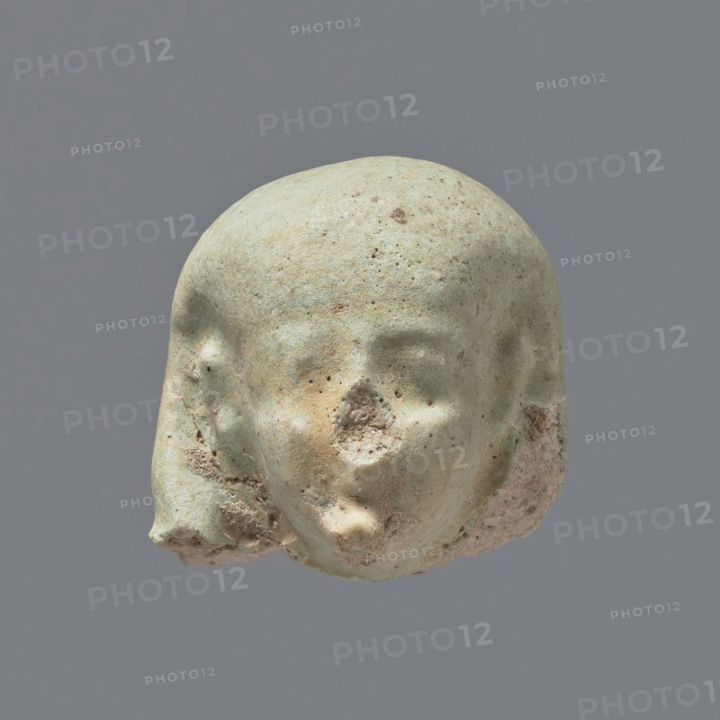 Fragment of a shabti. Head of a shabti figurine with not very carefully rendered facial features, in a smooth wig and with a thin beard not connected to the neck. Celadon glaze. 332–30 BC; Ptolemaic Period. El-Kantara. Shabti, height 2.5 cm., A small sculpted head fragment with a rounded top and stylized facial features, rendered in a pale greenish hue with areas of beige and brown discoloration; the surface shows pitting and rough texture, especially around the nose and lower jaw where some erosion has exposed a sandy, pinkish-beige interior; the left and right sides of the head have irregular chipped areas with a mix of green, beige, and pinkish tones; the background is a uniform medium gray.