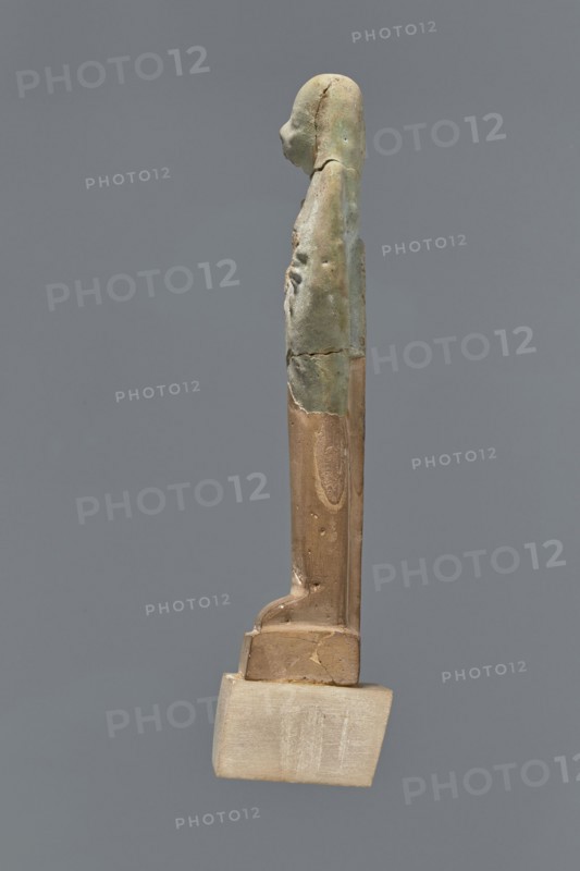Fragment of a shabti. Mummy-shaped figurine, grave good, preserved to the beginning of the thighs. Upper part of a shabti figurine with a smooth tripartite wig, holding hoes, and a back pillar; facial features are carelessly rendered. Celadon glaze. Ptolemaic Period, 332–30 BC; from El-Kantara. Shabti, height 7.8 cm., A slender vertical figurine viewed in profile against a solid gray background, with a rounded head and a tapered torso that shows surface irregularities and small dents; the upper portion of the figure has a greenish-blue patina with areas of lighter green and pale blue, while the lower portion is a warm brown with smoother texture and remnants of a lighter beige base at the bottom; overall the object shows seams and cracks where the colors meet and a subtle contrast between the cool green-blue and the warm brown and beige tones.