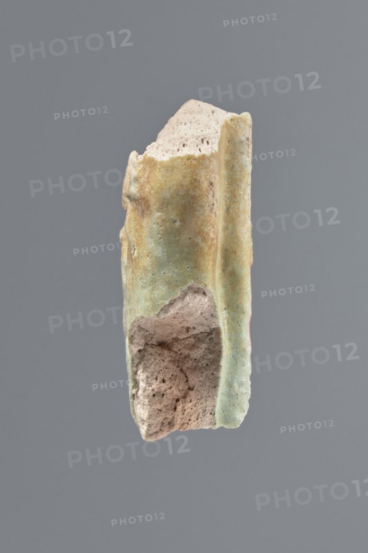Fragment of a shabti, unknown creator; mummy-shaped funerary figurine, grave goods. Middle part of the torso of a shabti figurine, preserved from the elbows to mid-thighs. Visible hands holding hoe tips, traces of an engraved column of inscription on the front, back pillar on the back. Celadon glaze. 332–30 BC; Ptolemaic Period, El-Kantara. Shabti, height 4.6 cm., A fragment of a porous object with an irregular, broken shape is centered against a solid gray background; the fragment shows rough, pitted interior surfaces in a light beige color and an outer surface with a weathered, greenish-tan patina, with areas where the beige interior is exposed and where the greenish-tan exterior varies in tone.