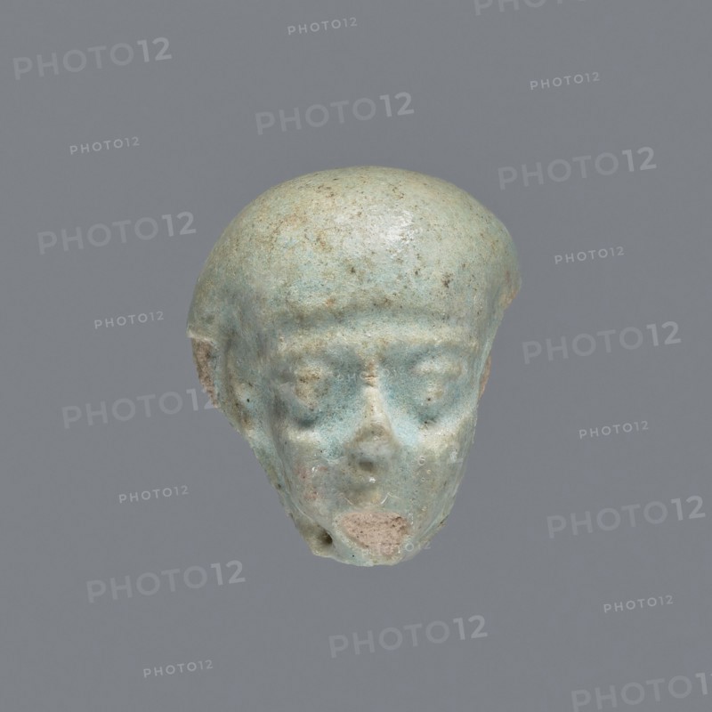 Fragment of a shabti; mummy-shaped funerary figurine. Head of a shabti figurine with carefully rendered facial features. Celadon glaze. Ptolemaic Period, 332–30 BC. From El-Kantara. Shabti; height 2.4 cm., A small sculpted head with a rounded top and simplified facial features, shown against a plain gray background; the object is primarily a pale bluish-green with areas of lighter and darker bluish tones and small patches of beige and brown, with a smoother sheen on the rounded scalp and a slightly rougher texture on the lower face, visible eye sockets, a subtle nose, and an eroded mouth area.