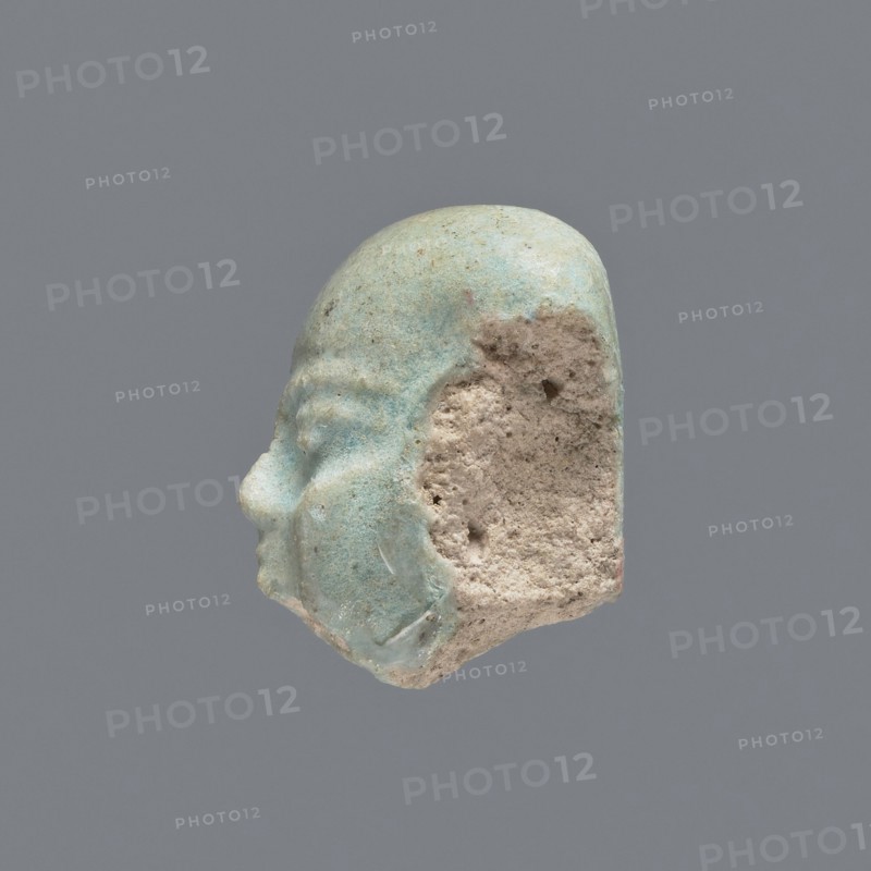 Fragment of a shabti;, Head of a shabti figurine with carefully rendered facial features. Celadon glaze. Mummy-shaped funerary figurine shabti. Ptolemaic Period, 332–30 BC. Found at El-Kantara. Height 2.4 cm., A small sculpted fragment of a human head shown in profile with visible facial features including a nose, closed eye, and rounded cheek, with a partially eroded rear section exposing a rough, porous interior; the outer surface is smooth and glazed in shades of pale blue and green with some areas of lighter cream and beige where the glaze is worn, while the exposed interior is a coarse, sandy beige; the background is a uniform gray.