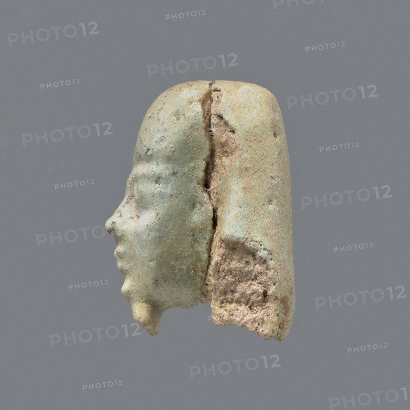 Fragment of a shabti. Head of a shabti figurine with carefully rendered facial features; smooth wig, thin beard. Celadon glaze. 332–30 BC; Ptolemaic Period. El-Kantara. Shabti; height 2.9 cm., A weathered sculpted head fragment shown in profile with a smooth rounded top and a separated rear section, exhibiting a pale green patina with areas of beige and light brown where the material is broken, a defined nose, lips, and chin on the face, a visible crack running vertically between the front and rear portions, and a few rough, porous surfaces on the broken edges against a neutral gray background.
