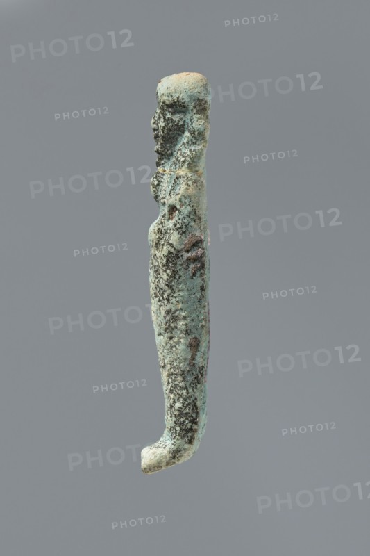Ushabti, mummy-shaped figurine. Flat back. Arms crossed on the chest, only the hands protruding from under the shroud, holding hoes in both hands. Tripartite wig on the head. Facial details illegible. Not inscribed. 664–332 BC Late Period, Memphis, Egypt. Height 8.5 cm., A vertically oriented elongated object with an irregular, slightly curved shape that resembles a narrow figure; its surface is rough and pitted with textured areas and small raised bumps. The upper portion has a rounded top and a suggestion of shallow contours on one side; the middle narrows slightly and then widens into a gently rounded lower section that curves outward at the very end. The color palette is predominantly greenish-blue with areas of darker green and black mottling, interspersed with patches of pale beige and subtle brown tones. The background is a uniform mid-gray.