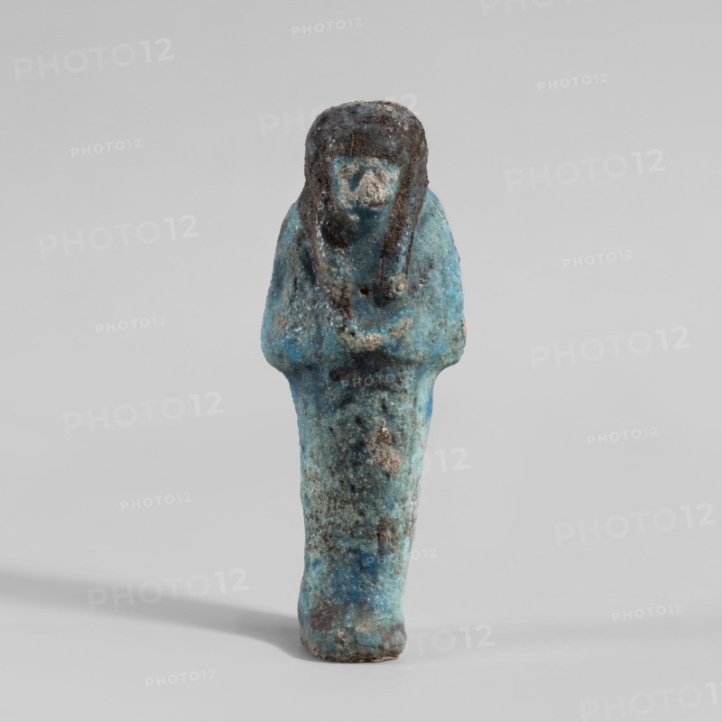 Ushabti, Mummy-shaped figurine. Hands folded on the chest—right over left. Hoes in each hand. Large basket slung over both shoulders on the back. Painted tools. Three-part smooth wig with a headband tied at the back; painted wig. Facial details blurred. Inscription engraved and filled with black dye on the front of the legs, now almost invisible. Third Intermediate Period, Deir el-Medina. Shabti. Height 10.7 cm, width 4 cm., A small statuette in shades of blue and turquoise with patches of darker blue and brown, shown upright on a light gray background; the figure has a rounded head with darker brown areas suggesting hair or a headdress, a simplified face with a lighter blue tone, narrow shoulders and arms folded across the chest, and a tapered lower body with mottled blue, turquoise, and brown surface texture and some rough, worn spots.