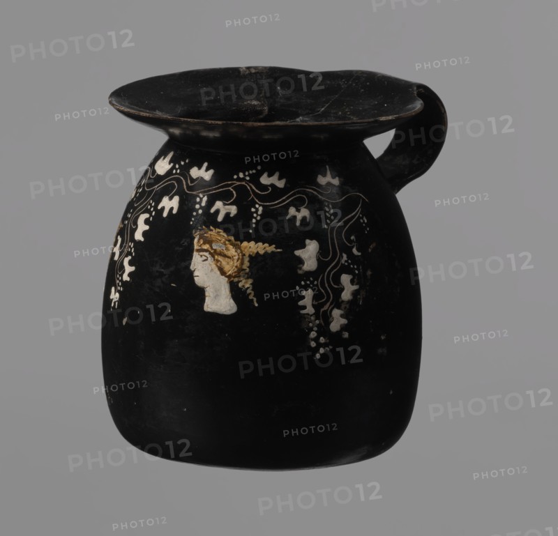 Gnathia-style mug, Gnathia style, Apulia, Southern Italy; 4th–3rd century BC. Red clay with glossy varnish, white-painted decoration and yellow retouching. Flat bottom, convex belly tapering at the top into a wide spout; band-like rim. Decoration on upper belly opposite the rim: three elongated female heads in profile facing left with large eyes and hairstyles shaded with yellow, surrounded by ivy fruits and leaves. Vessel/cup, antique Greek ceramics., A black ceramic vessel with a wide, flattened rim and a single loop handle showing signs of wear; a painted profile of a human head with golden-brown hair appears on one side, surrounded by decorative white motifs and wavy lines, with small white dot clusters and abstract white shapes cascading nearby; the background is gray.