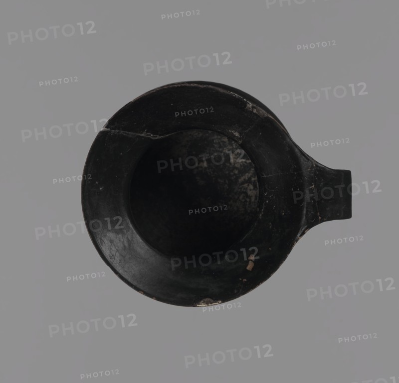 Gnathia-style mug, Antique vessel mug from Apulia, Southern Italy; 4th–3rd century BC. Red clay with glossy varnish, white-painted decoration and yellow retouching. Flat bottom, convex belly tapering to a wide spout; band-like rim. Decoration mainly on the upper belly opposite the rim: three elongated female heads in profile turned left with large eyes and hairstyles shaded with yellow, surrounded by an ornament of ivy fruit and leaves. Gnathia style; Greek/Ancient Greek pottery., A top-down view of a round vessel with a spout, the vessel appearing predominantly black with worn areas showing muted brown and beige tones, placed against a uniform gray background; the interior is dark with mottled lighter patches, and the rim shows a few small chips and scratches.