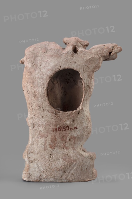 Figurine of Eros with a lyre, Eros myth., Greek deities, lyres musical instruments. Figurine of Eros standing on a base, holding a lyre resting on a base. 2nd century BC, Southern Italy . Terracotta figurine, height 19 cm, width 13.5 cm, depth 4.5 cm, weight 0.38 kg., A weathered sculptural object resembling a hollow form with an irregular rounded opening in its front, showing a rough, textured surface; the piece has an overall dusty pinkish-beige color with areas of light gray and pale brown, and a slightly darker interior visible through the opening; its silhouette includes a protruding, snout-like extension on one side and a small knob-like projection on the opposite side, with uneven contours, pits, and abrasions across the surface; the background is a flat mid-gray and the object rests on a smooth gray plane.
