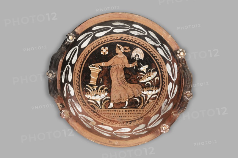 Red-figure patera-phiale depicting a woman with a fan and basket. Classified Gallo-Classical Scenography, female images iconographic. Southern Italian pottery. Repossession Zabkowice Slaskie. Apulia Italy. Reddish clay with black glaze; numerous additions of white and yellow paint. Deep bowl with two vertical, profiled handles on the rim, decorated with three decorative knobs with heads adorned with white rosettes. Funnel-shaped foot with an upturned, profiled rim; similar profiling on the vessel rim. Richly decorated interior: in a medallion framed by laurel-branch motif painted in white and a black chasing-wave ornament, a woman running left in a chiton holding a basket and a fan. Stylized plants on the sides; a rosette in the left field. Dated 350–325 BC previously 3rd quarter of the 4th century BC. Type: vessel, patera. Weight: 1.144 kg., A round decorative plate with a central painted scene featuring a standing woman in a flowing dress holding an object in each hand, surrounded by...