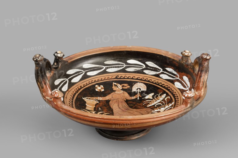 Red-figure patera-phiale depicting a woman with a fan and basket, Classified Gallo-Classical Scenography, female images iconographic. Southern Italian pottery; Apulia Italy. Reddish clay with black firnis; numerous additions of white and yellow paint. A deep bowl with two vertical, profiled handles on the rim, decorated with three decorative knobs with heads adorned with white rosettes. A funnel-shaped foot with an upturned, profiled rim; similar profiling on the rim of the vessel. Richly decorated interior: in a medallion framed with a motif of laurel branches painted in white and an ornament of black chasing waves, a woman running to the left in a chiton with a basket and a fan. On the sides are stylized plants; in the left field a rosette. Dated 350–325 BC previously 3rd quarter of the 4th century BC. Vessel type: patera. Weight 1.144 kg., A shallow ceramic bowl with a rounded foot and a wide rim, painted mainly in black and terracotta with white decorative elements, features a cent...