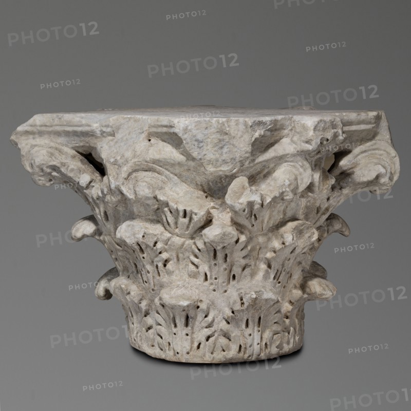 Corinthian capital, Sacred architecture, Roman architecture; capitals; acanthus ornament; rosettes. Corinthian capital similar to kalathos capitals. 3 rows of leaves. All leaves similar, with a wavy surface, divided by deep, softly bending grooves—veins—to the sides, rounded, with edges divided into lobes. The upper edge of the kalathos is visible above the leaves. An abacus with extended corners is composed of two plates of almost equal height. A round hole on the upper surface of the capital—probably a trace of contemporary mounting of the capital. A similar hole in the base. Great softness of modeling of details. Flakes separated by holes. 2nd century, Roman Empire 27 BC–476. Architectural element; element of architectural decoration; capital; architectural detail. Weight 133.5 kg., A carved stone architectural capital with a weathered surface, showing a group of ornate acanthus-like leaves and scrolls arranged around its cylindrical base and flaring top, with visible chips and roug...