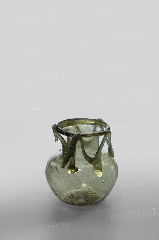 Flask, unknown eastern workshop, Scene: Gallic/classical? Scen.Gal.Klass., glass vessel, miniature, Type: Barag type VI:9. Transparent yellowish-green glass with applied thread decoration between the arms and the rim edge forming 10 sharp bends; free-blown. Outer rim diameter 5.50 cm; bottom diameter 3.90 cm. 4th century, Roman Empire 27 BC–476. Vessel, weight 0.076 kg., A small translucent glass vessel sits on a light surface against a pale background, the glass showing a greenish tint with areas of yellow-green on the applied rim and decorative drips; the body is rounded and clear enough to reveal internal bubbles and small inclusions, and the decorative strands hang from the rim in looping curves with thicker teardrop ends, all rendered in shades of green, yellow-green, pale gray, and subtle iridescent hints.