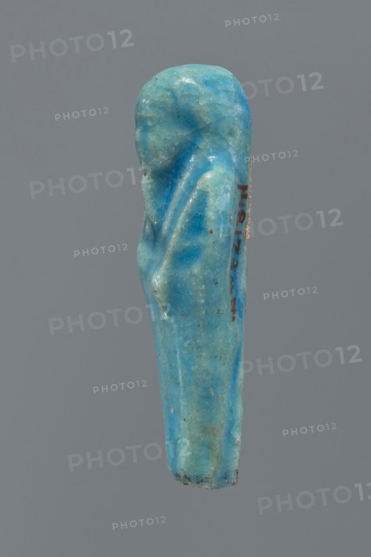 Shabti, mummy-shaped figurine, funerary gift; back supported by a pillar/pilaster. Legs missing up to knee height. Arms crossed on the chest, right folded over left. In the right hand is a hoe, in the left a pickaxe. On the head is a three-part, smooth wig. The beard is long, straight, and braided. Facial details are poorly visible. Not inscribed. 664–332 BC, Late Period, Egypt. Height 6.9 cm., A small carved object shaped like a standing figure with a rounded head and draped body, rendered in a smooth blue-green material with variations of turquoise and aquamarine tones, showing areas of lighter and darker blue-green, some pale greenish tints, and tiny dark specks and weathered spots; the surface has a slightly glossy, worn appearance with subtle ridges and folds suggesting drapery, and the object is photographed against a plain gray background.