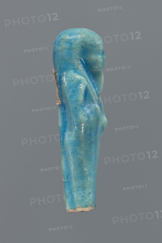 Shabti, Mummy-shaped figurine, back supported by a pillar/pilaster. Legs missing up to knee height. Arms crossed on the chest, right folded over left. In the right hand is a hoe, in the left a pickaxe. On the head is a three-part, smooth wig. The beard is long, straight, and braided. Facial details are poorly visible. Not inscribed. 664–332 BC, Late Period, Egypt. Height 6.9 cm., A small sculpted figurine shaped like a standing person shown in profile, rendered in a bright turquoise blue with lighter and darker blue variations and a few tiny dark specks, with a slightly glossy surface and visible cracks and wear; the figure has a rounded head, a subtle suggestion of a shoulder and arm, and a tapered body ending in a rough beige edge, all set against a plain medium-gray background.