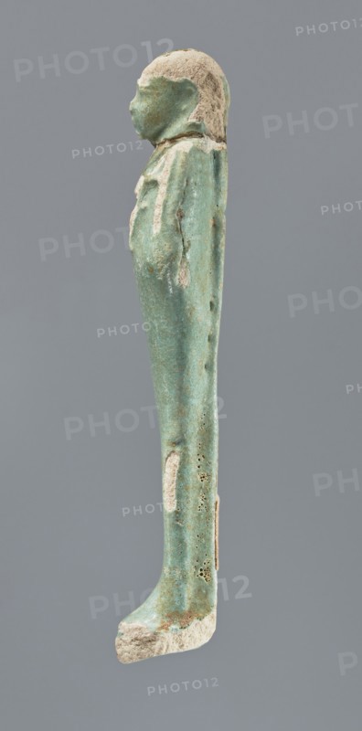 Shabti, mummy-shaped figurine, funerary gift; standing on a base with back supported by a pillar/pilaster. Arms crossed on the chest with only the hands protruding from under the shroud. Right hand holds a hoe and left hand a pickaxe, both tool ends wrapped. A basket is on the back, slung on a single twisted cord over the left shoulder. Wears a three-part smooth wig that merges into the pillar; long braided beard. Facial details poorly visible. Not inscribed. 664–332 BC, Late Period, Egypt. Height 12.5 cm., A narrow vertical statuette of a standing human figure photographed in profile against a plain gray background, with a rounded head and simplified facial features, a long wrapped body forming a straight column, and visible surface wear and erosion; the object shows a predominant turquoise-green color with patches of off-white and beige where the surface is worn, and small dark pitted areas on the side.