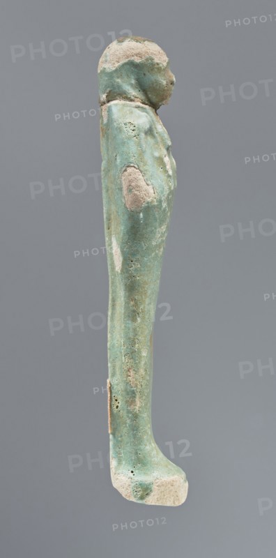 Shabti, mummy-shaped figurine, funerary gift, hoes; standing on a base, back supported by a pillar/pilaster. Arms crossed on the chest with only the hands protruding from under the shroud. Right hand holds a hoe, left a pickaxe; the ends of both tool handles are wrapped. A basket is on the back, slung on a single twisted string over the left shoulder. Wears a three-part smooth wig that merges into the pillar and a long braided beard. Facial details are poorly visible. Not inscribed. 664–332 BC, Late Period, Egypt. Height 12.5 cm., A narrow vertical turquoise-green object with areas of pale beige and off-white erosion, shaped like a standing human figure viewed in profile, showing a rounded head, a draped upper body, an elongated straight lower body and a flat foot; the surface has a weathered, pitted texture with several rough patches and small holes, and the background is a uniform cool gray.