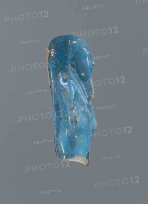 Shabti, Mummy-shaped figurine, funerary offering. Supported on a pillar at the back. Legs missing. Arms crossed on the chest, right folded over left. In the right hand is a hoe, in the left a pickaxe. On the back is a bag slung on a string over the left shoulder. Convex tools. On the head is a long, smooth, tripartite wig. Long, straight braided beard. Not inscribed. 664–332 BC; Late Period, Egypt. Shabti, height 5.2 cm., A small sculpted fragment of a human profile rendered in a glossy blue material with areas of lighter and darker blue and a few greenish spots, showing a head and upper torso with a smooth rounded head, a subtle nose, and a draped shoulder and chest, with the back edge revealing a beige, unglazed interior; the background is uniform gray.