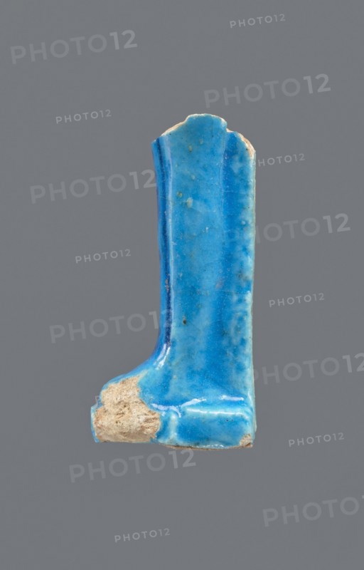 Shabti legs, Fragment of the legs of a mummy-shaped shabti figurine preserved from the knees down. The feet rest on a base, the back of the figurine supported by a pilaster. On the front of the legs is a painted hieroglyphic inscription in vertical lines. Mummy-shaped figurine, funerary gift; Late Period, Egypt, 664–332 BC. Shabti; height 8.5 cm, width 2 cm., A fragment of a glazed object with a glossy sky blue surface showing variations to deeper turquoise and small speckles, with a rough exposed beige-brown unglazed area at one end, overall L-shaped with a rounded top edge and a vertical ridge along one side, set against a plain neutral gray background.