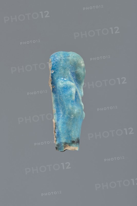 Shabti, Mummy-shaped figurine, funerary gift; supported on a pillar at the back. Legs missing. Arms crossed on the chest, right folded over left. In the right hand, a hoe; in the left, a pickaxe. On the back, a bag slung on a string over the left shoulder. Convex tools. On the head, a long, smooth, three-part wig. Long, straight beard braided. Facial details poorly visible. Not inscribed. 664–332 BC, Late Period, Egypt. Height 5.4 cm., A small, worn vertical object with an irregular rounded top and uneven surface in shades of bright blue, turquoise, and teal, with areas of lighter aqua and pale blue, and a few darker blue spots; the edges show rough beige and tan material with small patches of dark gray or black near the bottom; it is set against a flat medium-gray background.