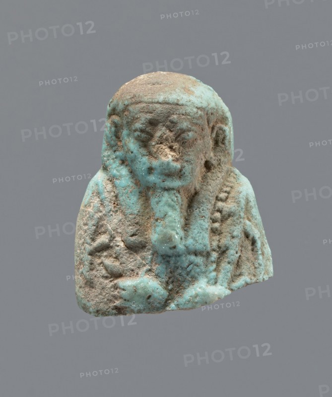 Ushabti - upper part of a figurine. Mummy-shaped funerary figurine ushabti, head and torso fragment. Figurine resting on a pillar with arms crossed on the chest, hoes in both hands. A twisted rope runs from the right hand over the left shoulder to the back. Convex tools; a basket hangs on the back on a string; the basket is engraved. Three-part smooth wig, long wide smooth beard. Large almond-shaped eyes, large ears; other facial details damaged. Inscription placed in a column on the pillar, text illegible. 664–332 BC, Late Period, Egypt. Height 3.5 cm., A small sculpted bust with a rough, weathered surface showing a mix of turquoise blue and sandy beige colors, depicting a rounded head with a stylized headdress or hair framing the face, a worn facial area with shallow eye and mouth forms, a vertical element at the front resembling a braided or folded feature, textured areas on the shoulders and chest with traces of turquoise glaze, and small patches of exposed beige material where the...