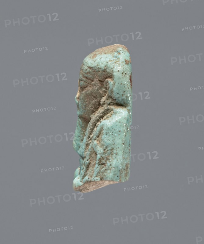 Ushabti - upper part of a figurine. Mummy-shaped funerary figurine, head and torso fragment. Figurine resting on a pillar with arms crossed on the chest, hoes in both hands. A twisted rope from the right hand runs over the left shoulder to the back. Convex tools; a basket hangs on the back on a string, the basket is engraved. Wearing a three-part smooth wig and a long, wide smooth beard. Large almond-shaped eyes and large ears; other facial details damaged. Inscription placed in a column on the pillar; text illegible. Late Period, Egypt, 664–332 BC. Ushabti; height 3.5 cm., A small turquoise-colored carved figure shown in profile against a plain gray background, with areas of brownish soil or patina on its surface and some lighter beige at the base; the figure has a rounded head, a prominent nose, closed eyes, and carved details suggesting hair or a headdress and draped clothing, with a textured, pitted surface and variations of turquoise, brown, beige, and small darker speckles.
