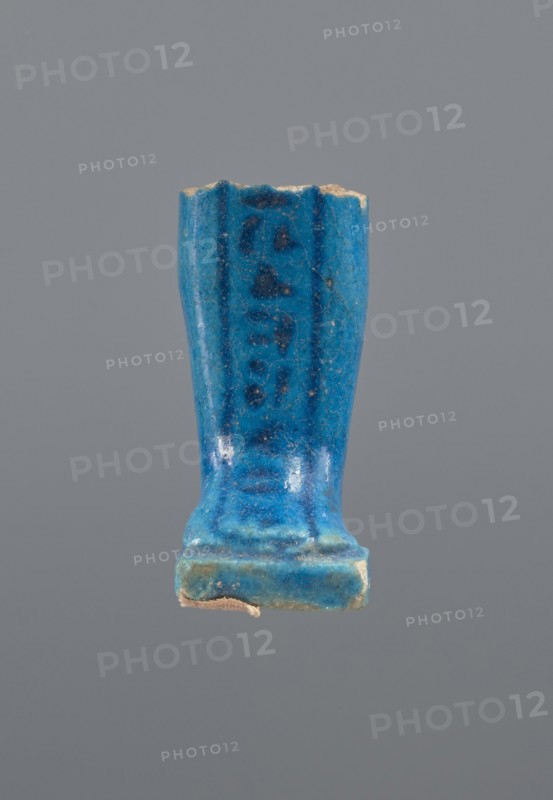Shabti legs, mummy-shaped figurine fragment: fragment of the legs of a mummy-shaped shabti figurine preserved from the knees down. The feet rest on a base, the back of the figurine is supported by a pilaster. In front of the legs is a column of hieroglyphic inscription enclosed in a frame; painted inscription. Late Period, Egypt, 664–332 BC. Shabti; height 4.3 cm, width 2 cm., A blue glazed ceramic or faience fragment shaped like a small upright rectangular column with a flared top and a stepped base sits against a plain gray background, showing worn edges and small chips where an underlying beige material is visible; the glaze displays variations of bright turquoise blue, deeper cobalt blue streaks, and some darker blue spots, with a few areas of lighter bluish-green near the base and tiny specks of pale beige from surface wear.