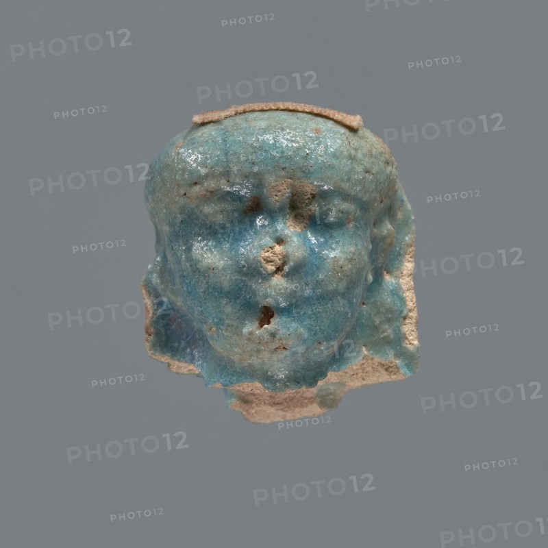 Shabti head, mummy-shaped figurine, funerary gift. Head of a shabti figurine depicting a king: oval face with full cheeks; prominent eyebrow arch; eye and nose shapes difficult to distinguish; corners of the mouth slightly upturned. Nemes placed low on the forehead, edges slightly outlined; end of the rear pillar visible at back of head. Ptolemaic Period, 332–30 BC, Egypt. Shabti; height 3 cm, width 2.3 cm., A small sculpted head fragment with a glossy turquoise-blue surface and rough beige areas where material is missing, showing the contours of a forehead, eye sockets, a nose area with chipped sections, and a partial mouth, with a flat beige strip across the top and uneven beige edges along the sides and bottom against a plain gray background.
