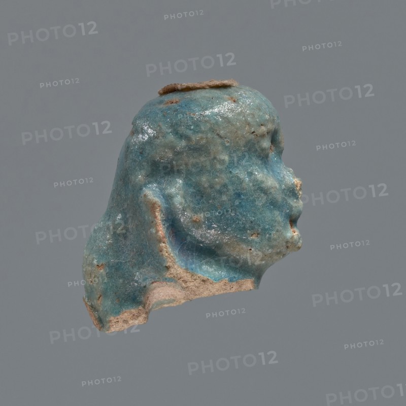 Shabti head, Mummy-shaped figurine head depicting a king. Oval face with full cheeks, prominent eyebrow arch; eye and nose shapes are indistinct. Corners of the mouth slightly upturned. Wears a low-set nemes whose edges are slightly outlined against the forehead. End of the rear pillar visible at the back of the head. Ptolemaic Period, 332–30 BC, Egypt. Shabti; height 3 cm, width 2.3 cm., A small sculpted fragment resembling a head in profile, with a weathered, glossy surface showing turquoise blue, teal, and pale blue-green tones mixed with areas of tan and beige where the material is exposed; the form has softened facial contours, a rounded top with a thin brown strip along the crown, and uneven, eroded edges revealing a rough, sandy interior against a uniform gray background.