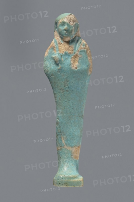 Ushabti. Mummy-shaped figurine standing on a base with a flat pillar at the back. Arms folded across the chest with delicately outlined hands visible from under the shroud; both hands hold hoes and a basket is slung over the left shoulder on the back. Wears a smooth, tripartite wig. Round, proportionate face. Not inscribed. 664–332 BC, Late Period, Egypt. Height 11 cm., A single small statuette shown against a plain gray background, with a turquoise-blue surface mottled with areas of beige where the coating has worn away; the figure has a rounded head with faint facial features and a suggestion of a headdress, a tapered body that widens slightly at the shoulders, a narrow lower section leading to a small flared base, and an overall textured, aged appearance with patches of turquoise and beige.