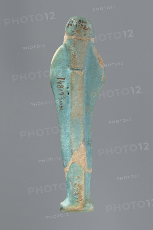 Ushabti, Mummy-shaped figurine standing on a base with a flat pillar at the back. Arms folded across the chest with delicately outlined hands visible from under the shroud; both hands hold hoes and a basket is slung over the left shoulder on the back. Wearing a smooth tripartite wig; round face with proportionate facial details. Not inscribed. 664–332 BC, Late Period, Egypt. Height: 11 cm., A slender figurine viewed from the back, primarily turquoise blue with areas of pale beige where the surface is worn, showing a narrow vertical ridge along the center back, rounded shoulders, a small rounded head, and a rectangular base; the surface has a slightly rough texture with some small pits and patches of darker blue and tan.