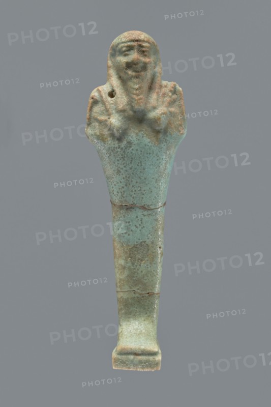 Ushabti, Mummy-shaped figurine shabti, 664–332 BC, Late Period, Egypt. Standing on a base with a flat pillar behind it. Arms folded across the chest with only the palms protruding from under the shroud; a hoe in the right hand and a pickaxe in the left, both tools convex with slightly curled handle ends. A twisted rope is slung over the left shoulder in the right hand, with a trapezoidal basket hanging from the rope on the back. Smooth three-part wig, long braided straight beard, round face with proportionate facial details. Not inscribed. Height 12.2 cm., A small elongated figurine with a rounded top resembling a human head and shoulders, a narrow tapered body and a flat rectangular base, showing a textured surface with pitting and areas of wear; the head and shoulders display simple facial features and a suggestion of raised arms across the chest, with a small hole visible near one shoulder; the object shows mottled colors of pale turquoise, greenish-blue, and patches of brown and be...