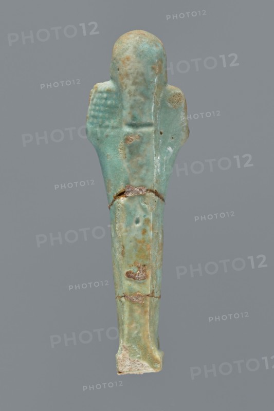 Ushabti, Mummy-shaped figurine shabti. Standing on a base with a flat pillar behind it. Arms folded across the chest with only the palms protruding from under the shroud. Hoe in the right hand and pickaxe in the left; ends of the handles slightly curled and tools rendered convex. A twisted rope slung over the left shoulder in the right hand, with a trapezoidal basket hanging from it on the back. Smooth three-part wig, long braided beard, round face with proportionate facial details. Not inscribed. Late Period, Egypt, 664–332 BC. Height 12.2 cm., A small elongated object shaped like a human figure, shown against a solid gray background, with a smooth rounded head and simplified arm shapes extending slightly from the sides; the surface is predominantly turquoise with areas of pale green, light blue, and patches of tan and brown discoloration, along with a few rough, worn spots and small chips revealing a lighter, off-white material at the base; the left arm area shows a textured grid pat...