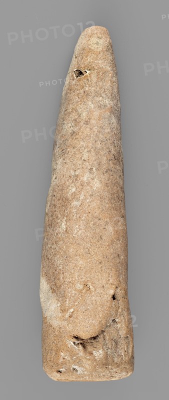 Mosaic cone, mosaic cones, decorative element, Uruk culture ca. 4000–ca. 3100 BC, Uruk Warka, Iraq, Sumer, 2nd half of 4th millennium BC, Southern Mesopotamia, Sumer, architectural decoration element, weight 44 g, A tall, elongated stone object with a smooth, tapered form displayed against a plain gray background; the stone shows a range of colors including light tan, sandy beige, warm brown, and patches of pale off-white, with several darker brown speckles and small rougher areas; its surface has subtle mottling and irregular textures, including a few small pits, shallow depressions, and rougher chips near the narrower end and along the wider base; overall the object appears rounded and slightly uneven in width with a natural, weathered appearance.
