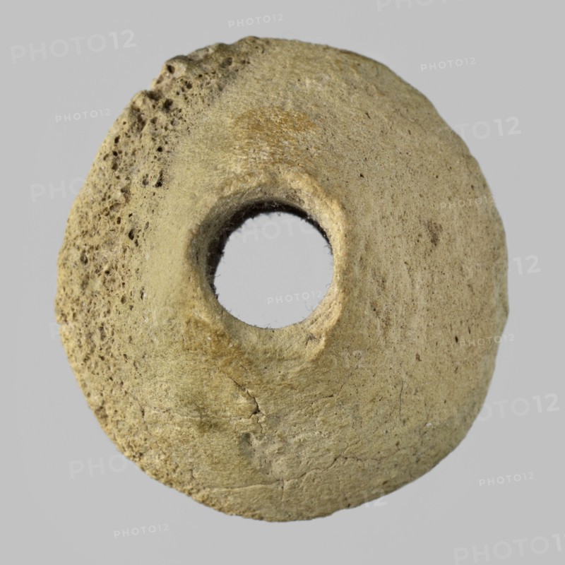 Spindle whorl, weaving tool, material culture, tools, Eastern Gaulish scene, 4th millennium BC, Sumer, Southern Mesopotamia, tool, weight 6.08 g, A roughly circular, doughnut-shaped object with a central hole, showing a textured, pitted surface on one side and a smoother surface on the other, with visible cracks and irregularities. The object appears in shades of beige, tan, and light brown against a light gray background.