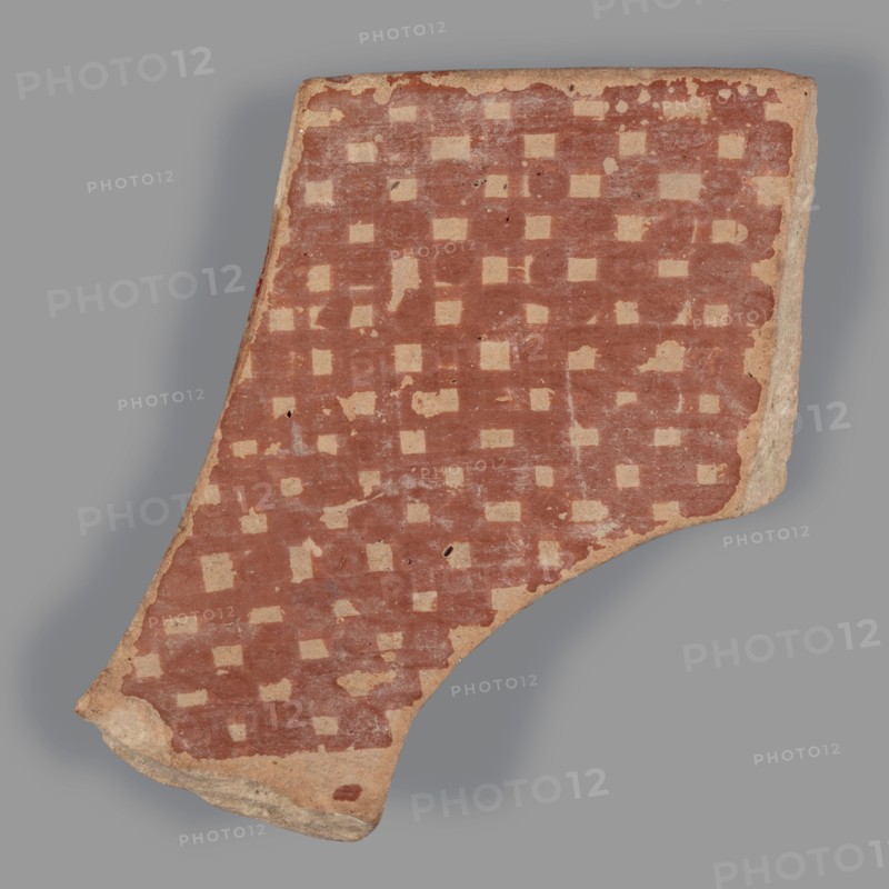 Fragment of a bowl, unknown Northern Mesopotamian workshop. Checkerboards, geometric motifs, painted pottery; Halaf culture c. 5200–c. 4500 BC; earlier dating c. 6500–c. 5500 BC. Northern Mesopotamia Iraq/Syria/Turkey. Pottery fragment: wall of a bowl with preserved thin rim. Before firing, decoration of geometric motifs was painted with ferrous clay on both sides over a layer of dried, heavily smoothed slip/glaze; after firing the painted motifs are red, background cream on the outside and light brown on the inside. Outer decoration: banded composition from top downward — a wide horizontal band, then alternating rows of single dots and two continuous thin lines, below which a single thin line with superimposed irregular ovals. Inner surface covered with a fine red checkered motif. Material: pottery. Dimensions: height 4.7 cm, width 4.6 cm, depth 0.5 cm; weight 10 g., A broken shard of pottery photographed against a gray background, showing a worn surface patterned with a grid of light...