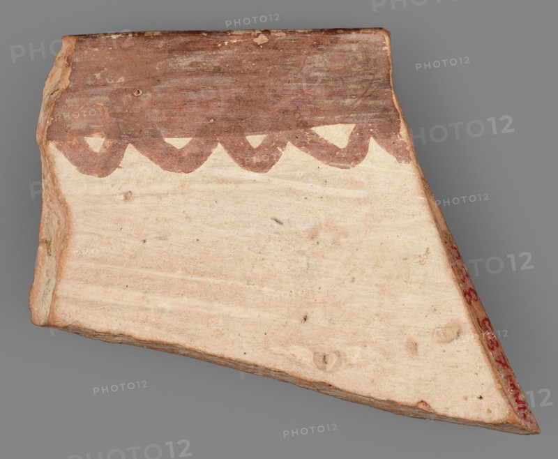 Fragment of a bowl, unknown Northern Mesopotamian workshop, painted pottery with geometric motifs, bowls, Eastern Gaulish Scenery. Fragment of the wall of a bowl or cup with a preserved pointed rim. On the outside, the decorative composition consists of from the top: a wide band of diagonally intersecting parallel lines, terminated at the bottom by a thick line. Below is a fragment of another band filled with a row of triangles or rhombuses filled with diagonally intersecting lines. On the inside, the decoration is limited only to the upper part of the vessel and consists of a wide band terminating at the bottom with a wavy line. c. 6500–c. 5500 BC; Halaf culture ca. 5200–ca. 4500 BC, Northern Mesopotamia Iraq/Syria/Turkey. Pottery fragment; height 3.8 cm, width 3.8 cm, depth 0.48 cm, weight 8 g., A fragment of a flat, triangular ceramic shard with a smooth, pale beige surface and a rougher, darker brown top edge that features a scalloped painted pattern; the shard shows worn, uneven e...