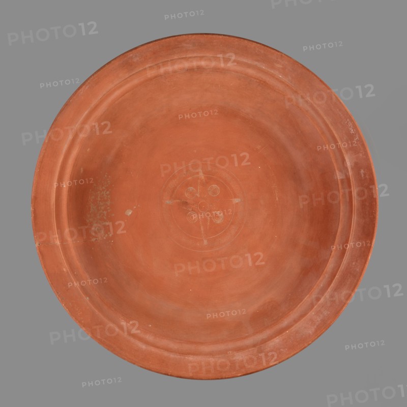 Bowl with palm leaf decoration. Roman pottery, Late Roman Red Slip Ware A Late Roman African pottery, ca. 360–450. Fine-grained clay, pink-brown 2.5YR 5/6; brown-red glaze 2.5YR 4/8–5/8 covering entire surface with slight sheen and slight cracking; inside shows slight glaze reflections. Thin-walled technique, smooth fracture Group A. In center of the bottom: stamped decoration forming a medallion — four palmettes and four triple rings in radial arrangement, surrounded by a roulette band between two concentric fluted grooves. Stamps: palmette Hayes type 10; triple rings Hayes type 26; stamped decoration classified as Palm Style B. Provenance: Carthage Tunisia, Africa Proconsularis. Vessel bowl weight 0.824 kg; height 5.9 cm; diameter 32.8 cm., A large round shallow dish viewed from above with a smooth terracotta surface in shades of reddish-brown and orange, showing a slightly raised rim and concentric circular indentations; the central area has a faint carved circular motif with a cros...