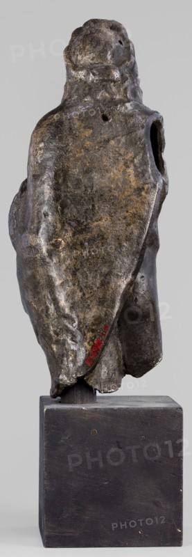 Figurine of Sylvanus in goat skin, with a pine wreath and a pine branch in his hand, figurines from a mold, Silvanus myth., 2nd century, Gaul France, figurine, height 23 cm, width 10 cm, depth 7.3 cm, weight 1.474 kg, A weathered sculptural form mounted on a rectangular block base, shown from the back. The sculpture has an irregular, organic surface with a draped, cloak-like outline and a rounded top suggesting a head. The surface shows varied textures with rough, pitted areas and smoother worn patches. Visible colors include dark brown, black, and patches of muted golden-brown and gray on the sculpture, while the base is a matte dark brown to black. There are a few small holes and indentations on the sculpture and an opening at one side, and the base shows subtle wear marks and small circular fastenings.