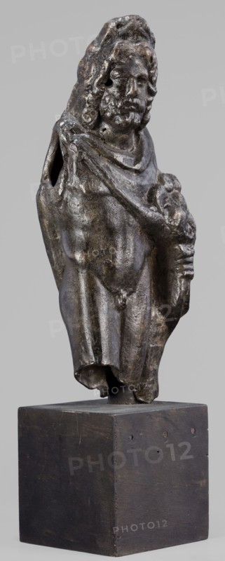Figurine of Sylvanus in goat skin, with a pine wreath and a pine branch in his hand, figurines from a mold, Silvanus myth., 2nd century, Gaul France, figurine; height 23 cm, width 10 cm, depth 7.3 cm, weight 1.474 kg, A small sculpted figure mounted on a rectangular base, shown full-length from front; the figure appears to be a standing male with long hair and a beard, wearing a draped cloak that covers one shoulder and falls to mid-thigh, with a rolled or folded section near the neckline; one arm is bent and holds a rounded object against the chest while the other arm is partly obscured by the cloak and has an open hollow at the shoulder; the surface of the figure has a dark metallic patina with areas of lighter worn highlights in brownish and gray tones and patches of darker almost black coloration; the base is a plain block with a dark brown to black finish and visible small marks and tiny holes on its sides; the background is a uniform light gray.