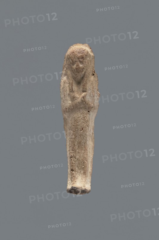 Shabti, mummy-shaped figurine, funerary gift, hoes; made carelessly, impressed in a mold. Hands folded on the chest, right over left; both hands hold hoes. Wears a three-part smooth wig. Facial details poorly legible. No inscription. Back unfinished. c. 1069–c. 656 BC, Third Intermediate Period, Egypt. Height 7.5 cm., A small elongated carved figure against a solid gray background, rendered in rough textured beige and light brown material with traces of darker brown in crevices; the figure has a rounded top suggesting a head with an indistinct facial area and a narrow tapering body, a subtle suggestion of crossed arms at chest level, and an uneven, slightly darker base; surface shows pitting and irregularities and a few small holes, with overall colors including beige, light brown, darker brown, and gray.