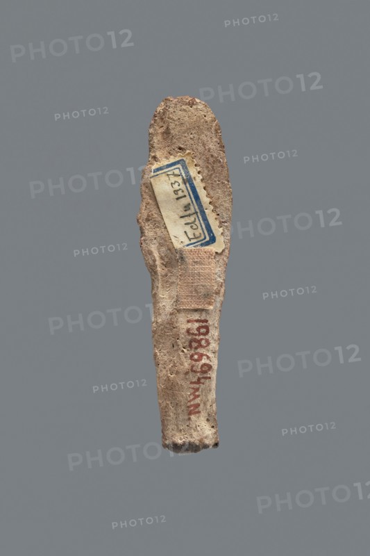 Shabti, Mummy-shaped figurine, funerary gift, made carelessly, impressed in a mold. Hands folded on the chest, right over left, both hands hold hoes. Wears a three-part smooth wig. Facial details poorly legible. No inscription. Back unfinished. c. 1069–656 BC; Third Intermediate Period, Egypt. Shabti, height 7.5 cm., An elongated, irregular object with a rough, porous surface in shades of beige, tan, and light brown is shown against a solid gray background; a small rectangular patch of woven fabric in a muted beige tone is attached near the midsection, and a rectangular paper label with a blue border and off-white interior is affixed near the upper portion, along with traces of reddish-brown markings on the lower area.